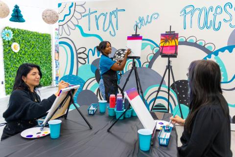 Two women painting on canvases in a Paint & Sip class at Ex Animo Creation with owner, Tatyanna Thomas, guiding the class.  