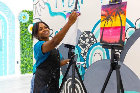 Woman smiling while painting a sunset-themed canvas on an easel at Ex Animo Creations in St. Cloud, Florida.