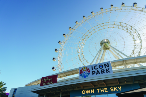 A low-angle view of The Wheel at ICON Park in Orlando, with clear blue sky overhead and signs for ICON Park and Madame Tussauds visible at the entrance.