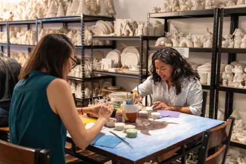 Two women painting ceramic pieces at a table surrounded by shelves of unpainted pottery inside Just a Glazin’ art studio.