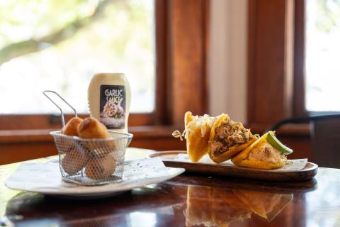 A plate of golden mini arepas filled with shredded pork, chicken, and cheese, served alongside crispy tequeños in a small fryer basket, with a bottle of garlic sauce in the background on a sunlit café table.