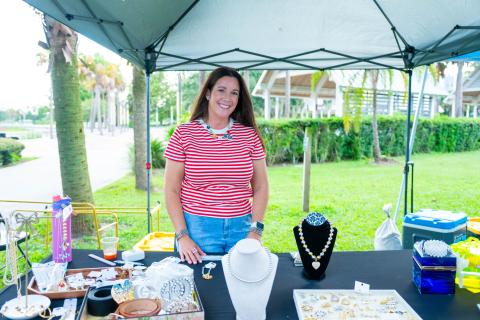 A vendor at the Kissimmee Valley Farmers Market smiles behind a jewelry booth, displaying necklaces, bracelets, and accessories at Lakefront Park.