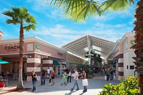 Shoppers stroll through an outdoor shopping plaza lined with palm trees and brand-name stores at Orlando Premium Outlets near Kissimmee, Florida.