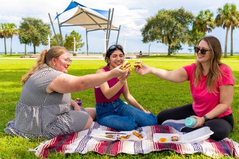 Three women enjoying a picnic on a blanket at Kissimmee Lakefront Park, toasting sandwiches together.