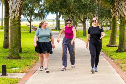 Three women exercising together at Kissimmee Lakefront Park, walking and roller-skating along a tree-lined path.