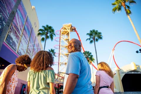 Family smiling and walking toward a towering red roller coaster under a bright blue sky at a theme park in Kissimmee, Florida.