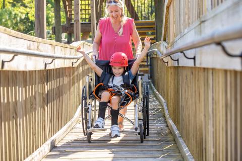 Child using a wheelchair with a zipline harness raises their arms in excitement while being assisted down an accessible wooden ramp at Gatorland in Kissimmee, Florida.