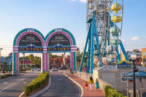 Entrance to Old Town Kissimmee, Florida, featuring the colorful archway and Ferris wheel against a bright blue sky, with visitors walking along the brick pathway.