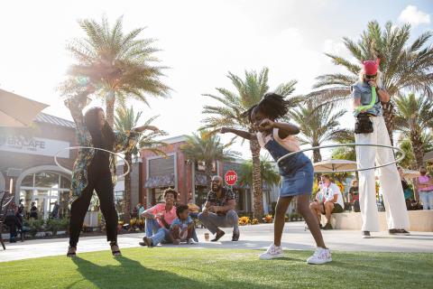 Families enjoying outdoor entertainment at Promenade at Sunset Walk in Kissimmee, Florida, with a woman and young girl hula-hooping under palm trees on a sunny afternoon.