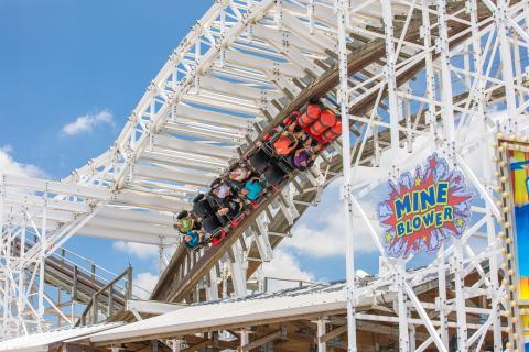 Riders hang upside down on the Mine Blower wooden roller coaster at Fun Spot Kissimmee, with the track twisting overhead against a bright blue sky.