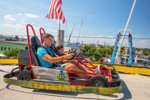 A father and daughter race side by side in go-karts at Fun Spot America Kissimmee, smiling as they speed around the elevated track under a sunny Florida sky.