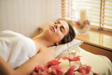 Woman relaxing on a massage table wrapped in a white towel, surrounded by soft lighting, flowers, and candles in a tranquil spa setting.