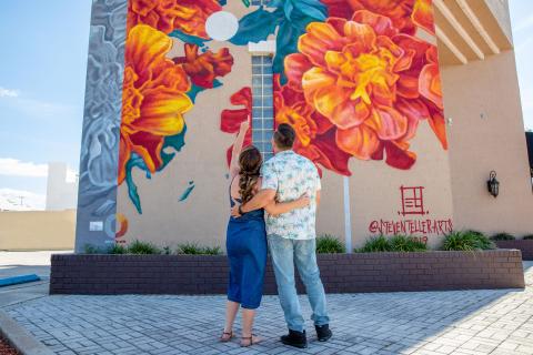 A couple stands arm in arm, looking up and pointing at a large, colorful floral mural painted on the side of a building in a sunny downtown setting.