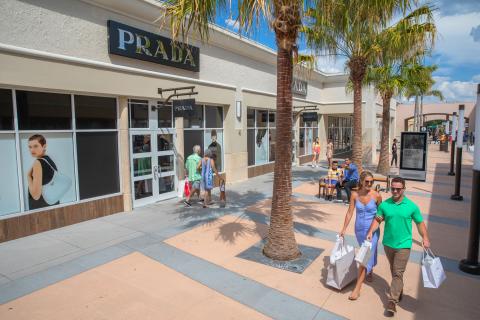 Shoppers stroll along a palm-lined outdoor shopping promenade, passing luxury storefronts while carrying branded shopping bags on a sunny day.