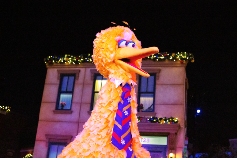 A large yellow bird character in a festive tie stands on an outdoor stage at night, with holiday lights and building façades glowing in the background.