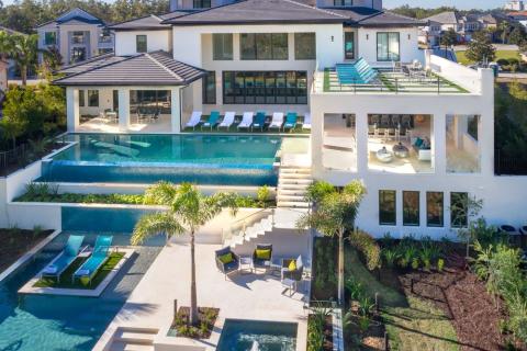 A large luxury vacation home in Kissimmee with multiple levels, expansive terraces, palm trees, and several pools including an infinity pool and a spa, viewed from above on a sunny day.