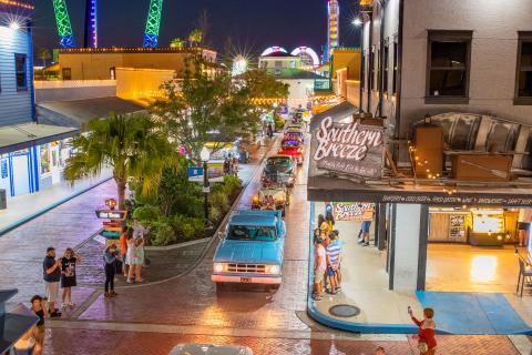 A nighttime classic car cruise through Old Town Kissimmee, with colorful neon lights, people watching from the sidewalks, and illuminated thrill rides glowing in the background.