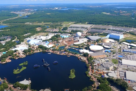 Aerial view of EPCOT at Walt Disney World Resort, showing Spaceship Earth, World Showcase Lagoon, surrounding pavilions, and nearby green spaces and roadways stretching into the distance.