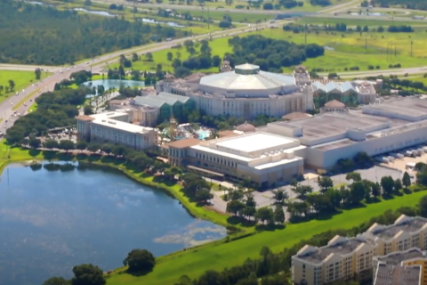 Aerial view of Gaylord Palms Resort in Kissimmee, showcasing its glass atriums, large event spaces, surrounding greenery, nearby roads, and a reflective lake in the foreground.