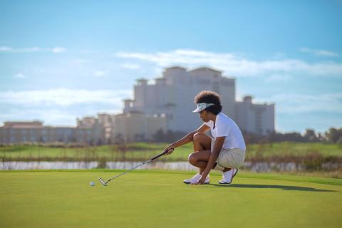 A woman kneels on the putting green of a golf course, aligning her shot with a putter as a large resort building stands blurred in the background.