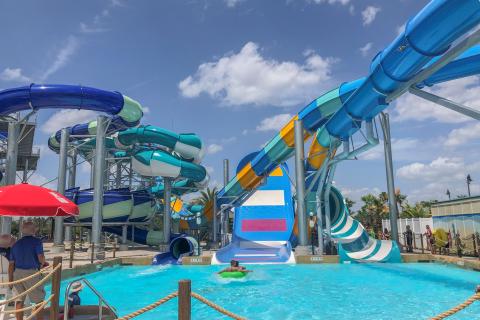 Colorful water slides twist and turn above a bright blue splash pool as a rider on a green tube glides into the water at an outdoor water park.