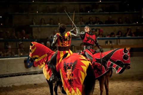 Two armored knights on horseback clash swords during a live performance at Medieval Times, with colorful heraldic costumes and an audience watching from tiered seating.  Caption (Gentle Joker):