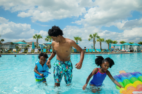 A father and two young children play together in a bright blue resort pool, splashing in the shallow water with palm trees, cabanas, and lounge chairs in the background.