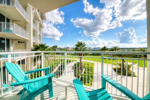 A private balcony with turquoise Adirondack chairs overlooking a sunny resort landscape with palm trees, walking paths, colorful buildings, and a distant lake under a bright blue sky.