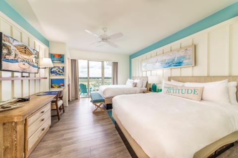 A bright, coastal-themed hotel room with two queen beds, colorful artwork, a desk, a mounted TV, and sliding glass doors leading to a balcony with outdoor seating and scenic views.
