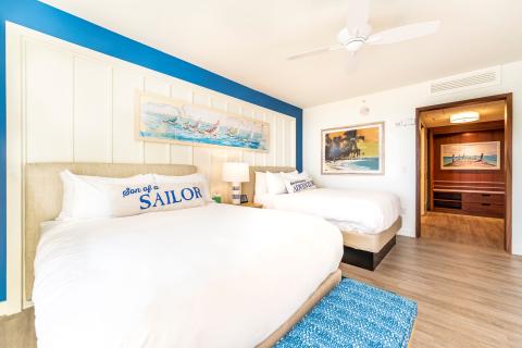 A bright, coastal-themed hotel room featuring two queen beds with white linens, nautical accent pillows, beach-inspired artwork, and warm wood finishes at Margaritaville Resort Orlando.