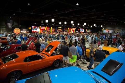 A bustling indoor Mecum Auctions event with a large crowd gathered around rows of classic cars, many with their hoods open, under bright overhead lights and colorful auction stage displays.
