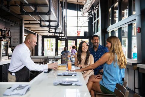A bartender serves drinks to three smiling guests seated at a modern bar with large windows, creating a bright and lively atmosphere.