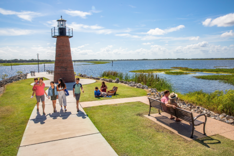Families enjoying a sunny day at Kissimmee Lakefront Park, walking near the brick lighthouse, relaxing on benches, and picnicking by the water.