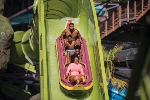 A family of five rides a multi-person raft down a steep green water slide at Universal’s Volcano Bay, splashing through water with excited expressions.
