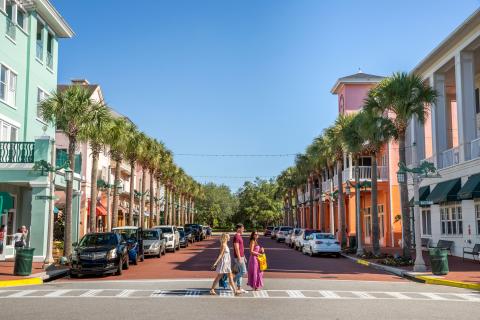 Friends crossing a palm-lined street in Celebration, Florida, surrounded by colorful pastel buildings, parked cars, and clear blue skies.