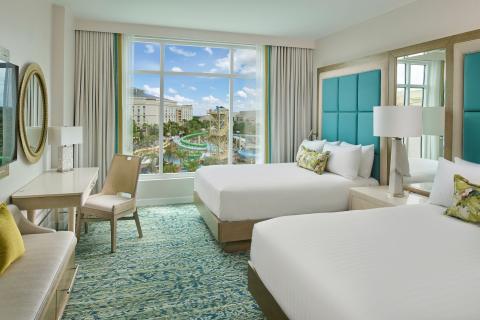 Bright, modern hotel room with two queen beds, teal headboards, a desk, and a large window overlooking the Gaylord Palms water park in Kissimmee, Florida.