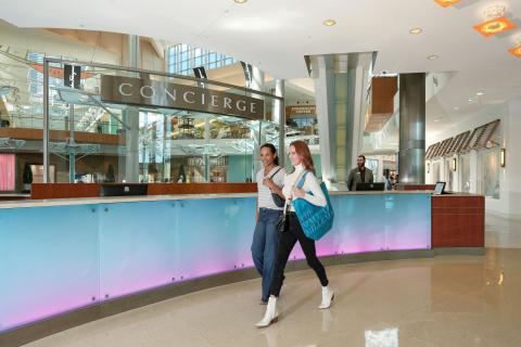 Two women walk past the concierge desk inside The Mall at Millenia, carrying shopping bags in a bright, modern interior with glass accents and high ceilings.