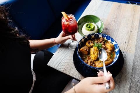 A diner sits with a bowl of posole and a glass of sangria on the table before her