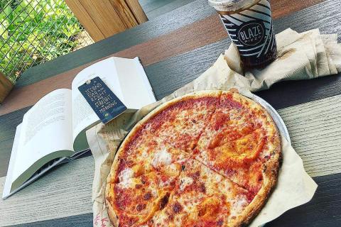 A freshly baked cheese pizza on a tray sits on a wooden picnic table beside an open book and an iced coffee from Blaze Pizza, creating a relaxed outdoor dining scene.