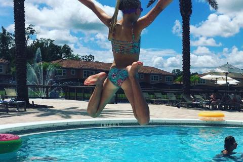A child leaps joyfully into a bright blue resort pool, arms stretched wide, framed by tall palm trees and a sunny Florida sky. Other swimmers relax in the water and on poolside loungers in the background.