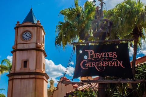 Entrance sign for the Pirates of the Caribbean attraction at Magic Kingdom, featuring a clock tower, palm trees, and a pirate ship mast under a bright blue sky.