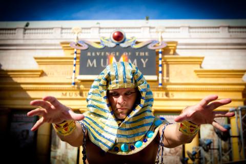 A man wearing a pharaoh costume stands in front of Revenge of the Mummy at Universal Studios and reaches out toward the camera with both arms.