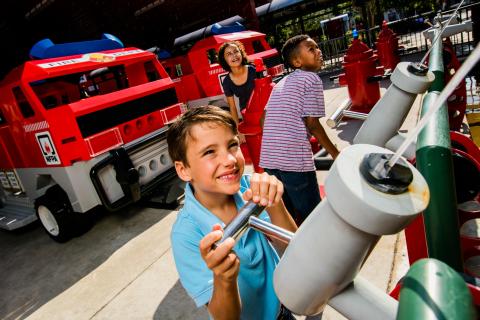 Children play at the Rescue Academy attraction at LEGOLAND® Florida Resort, spraying water from hoses toward LEGO fire trucks in a hands-on firefighting adventure.