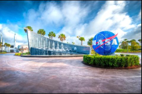 Entrance to Kennedy Space Center Visitor Complex in Merritt Island, Florida, featuring the iconic blue NASA globe and the fountain-lined Space Mirror Memorial under a bright blue sky.