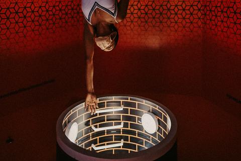 A person appears to be suspended upside down inside the Museum of Illusions, reaching between two circular openings in a dramatic optical illusion room with red geometric walls.