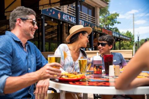 A family enjoying an outdoor meal together at a restaurant patio, smiling and talking over drinks and plates of food on a sunny day.
