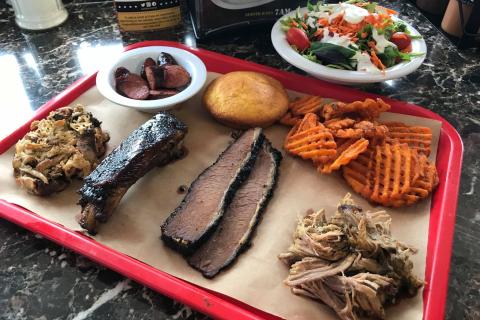A red tray filled with barbecue items including brisket slices, ribs, pulled pork, smoked sausage, sweet potato waffle fries, and a cornbread muffin, with a side salad and salad dressing in the background.