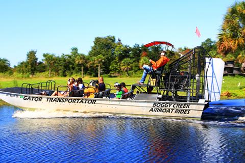 An airboat full of passengers glides across the water at Boggy Creek Airboat Rides, surrounded by bright greenery and blue skies.