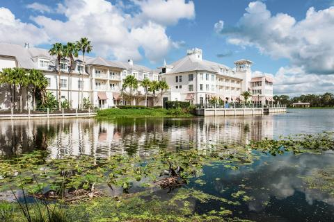 Waterfront view of The Inn at Celebration, with pastel-colored buildings, palm trees, and their reflections mirrored in a calm lake dotted with lily pads under a partly cloudy blue sky.