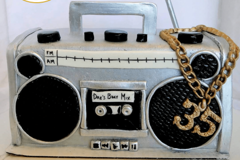 Custom boombox-shaped cake with silver and black detailing, a radio dial labeled FM/AM, a cassette reading “Dre’s Bday Mix,” and a gold chain with a dollar-sign pendant, created by Candyland Designs Co.